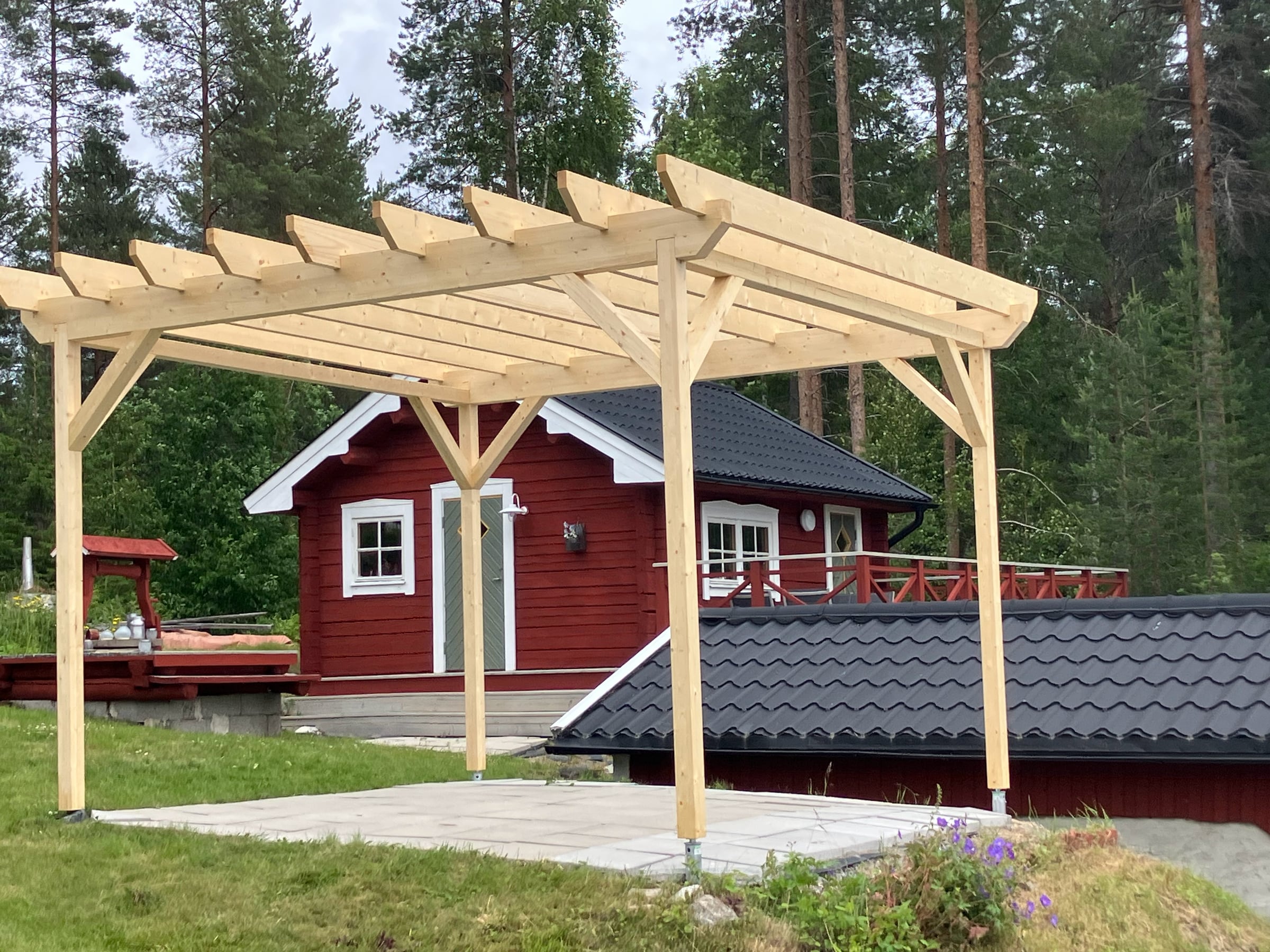 Nybyggd pergola i trä på en stenlagd uteplats framför en röd stuga